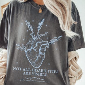 May include: Dark gray t-shirt with a light blue anatomical heart graphic and the text "NOT ALL DISABILITIES ARE VISIBLE" and "pete syndrome awareness". The shirt is a comfortable, casual style.