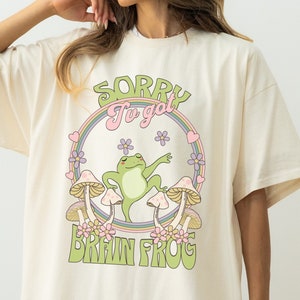 Puede incluir: Una camiseta blanca de gran tamaño con un estampado gráfico de una rana verde bailando sobre setas con el texto "Sorry I've got Brain Frog" en un círculo arcoíris.