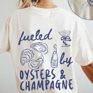 Puede incluir: Camiseta color crema con un gráfico azul marino en la espalda que dice "Fueled by Oysters & Champagne". El diseño incluye ilustraciones de ostras, una botella de champán y una copa de champán. Una versión más pequeña del diseño está en el frente.