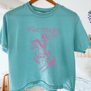May include: A turquoise t-shirt with the text "Y'all Means All" in pink, along with a pink graphic of a person riding a horse. The shirt is hanging on a wooden hanger.