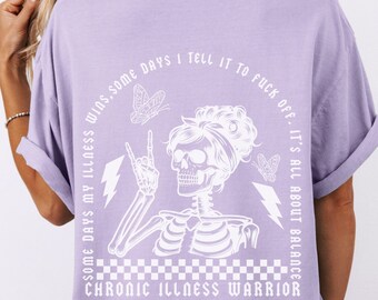 May include: Purple t-shirt with a white graphic of a skeleton with a crown of flowers, holding up a peace sign. The graphic also includes butterflies and lightning bolts. The text on the shirt reads "Some days my illness wins, some days I tell it to fuck off. It's all about balance. Chronic Illness Warrior."