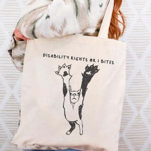 Könnte beinhalten: Cremefarbene Tragetasche mit einer schwarz-weißen Katzenillustration. Die Katze ist mit erhobenen Pfoten dargestellt, und der Text "DISABILITY RIGHTS OR I BITES" ist darüber gedruckt. Die Tasche hat ein schlichtes, minimalistisches Design.