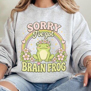 Pode incluir: Uma camisola cinzenta clara de gola redonda com uma impressão gráfica de um sapo verde a usar uma coroa de flores. O design inclui um arco-íris pastel, flores cor-de-rosa e o texto SORRY I've got BRAIN FROG em letras verdes e rosas.