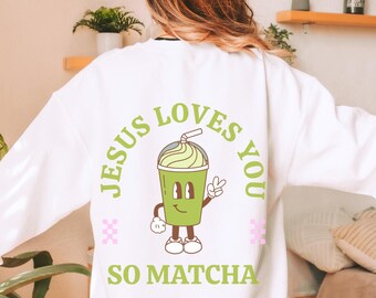 May include: White sweatshirt with a green cartoon matcha drink character and the text "Jesus Loves You So Matcha".