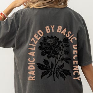 May include: Dark gray t-shirt with a black floral design and the text "RADICALIZED BY BASIC DECENCY" in a peach color. The shirt is a relaxed fit and has short sleeves.
