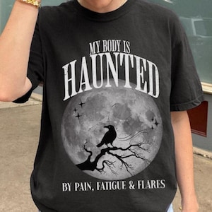 May include: Black t-shirt with the text "MY BODY IS HAUNTED" in white, above a graphic of a full moon, a crow on a branch, and the text "BY PAIN, FATIGUE & FLARES". The shirt is worn by a person.