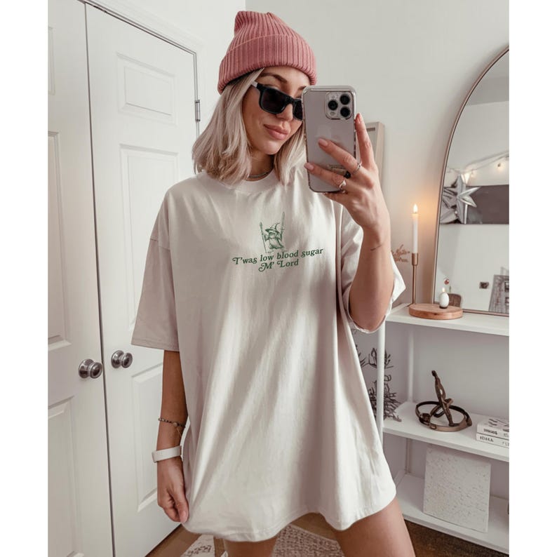 May include: Oversized beige t-shirt with the text "T'was low blood sugar M'Lord" in green. The shirt is paired with a pink beanie and black sunglasses. The person is holding a phone and standing in front of a mirror.