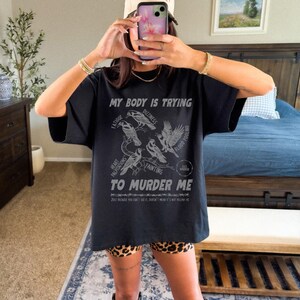 Pu&ograve; includere: T-shirt nero con grafica con la scritta "MY BODY IS TRYING TO MURDER ME" e illustrazioni di uccelli. La maglietta elenca anche sintomi come affaticamento, vertigini e svenimenti. La parte inferiore della maglietta riporta la scritta "JUST BECAUSE YOU CAN'T SEE IT, DOESN'T MEAN IT'S NOT KILLING ME."