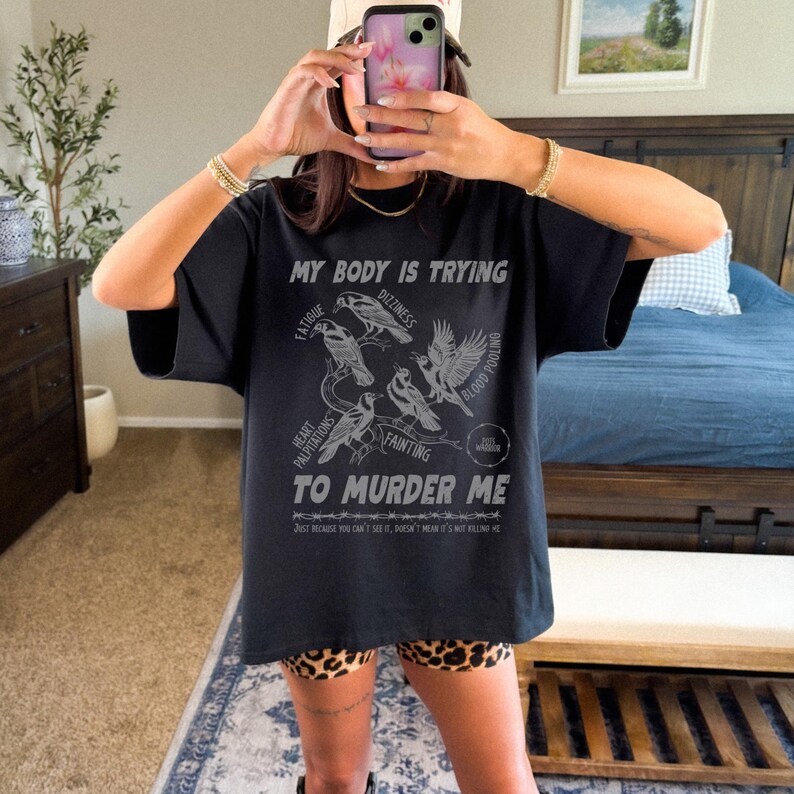 Pu&ograve; includere: T-shirt nero con grafica con la scritta "MY BODY IS TRYING TO MURDER ME" e illustrazioni di uccelli. La maglietta elenca anche sintomi come affaticamento, vertigini e svenimenti. La parte inferiore della maglietta riporta la scritta "JUST BECAUSE YOU CAN'T SEE IT, DOESN'T MEAN IT'S NOT KILLING ME."