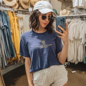 May include: A navy blue t-shirt with the text "T'was low blood sugar M'Lord" in gold script. The shirt is paired with light-colored checkered shorts. The person is wearing a white baseball cap and sunglasses.