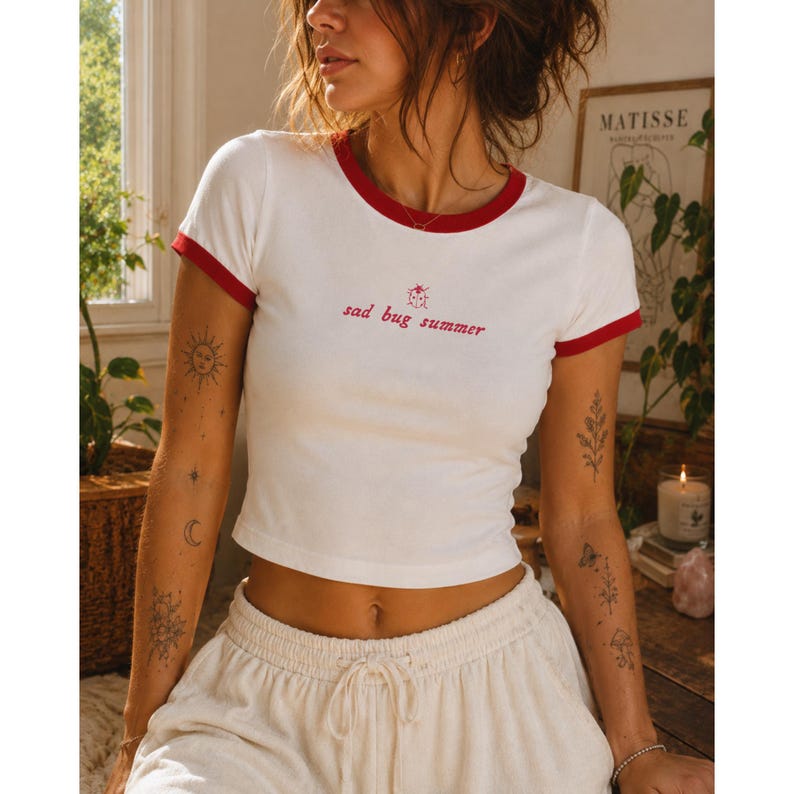 May include: A white ringer t-shirt with red trim on the collar and sleeves. The chest features a small red ladybug graphic above the red text that reads "sad bug summer" in a cursive font. The shirt has a cropped fit and short sleeves.
