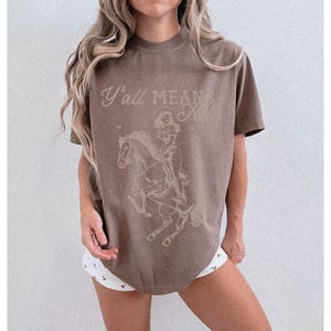 May include: A brown t-shirt with the text "Y'all Mean All" above a graphic of a skeleton riding a horse. The design is in a lighter brown color. The shirt has a crew neck and short sleeves.