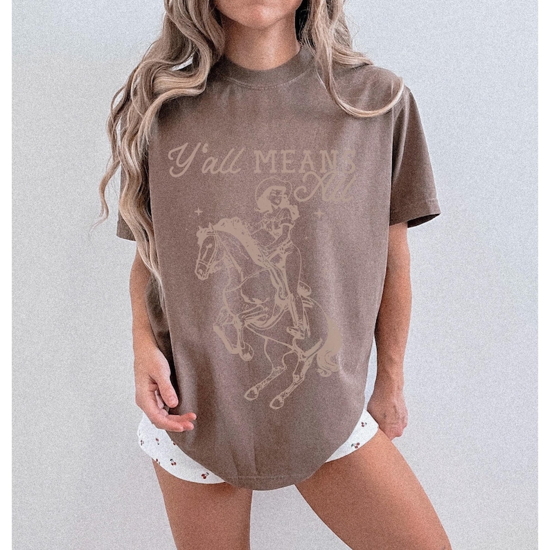 May include: A brown t-shirt with the text "Y'all Mean All" above a graphic of a skeleton riding a horse. The design is in a lighter brown color. The shirt has a crew neck and short sleeves.