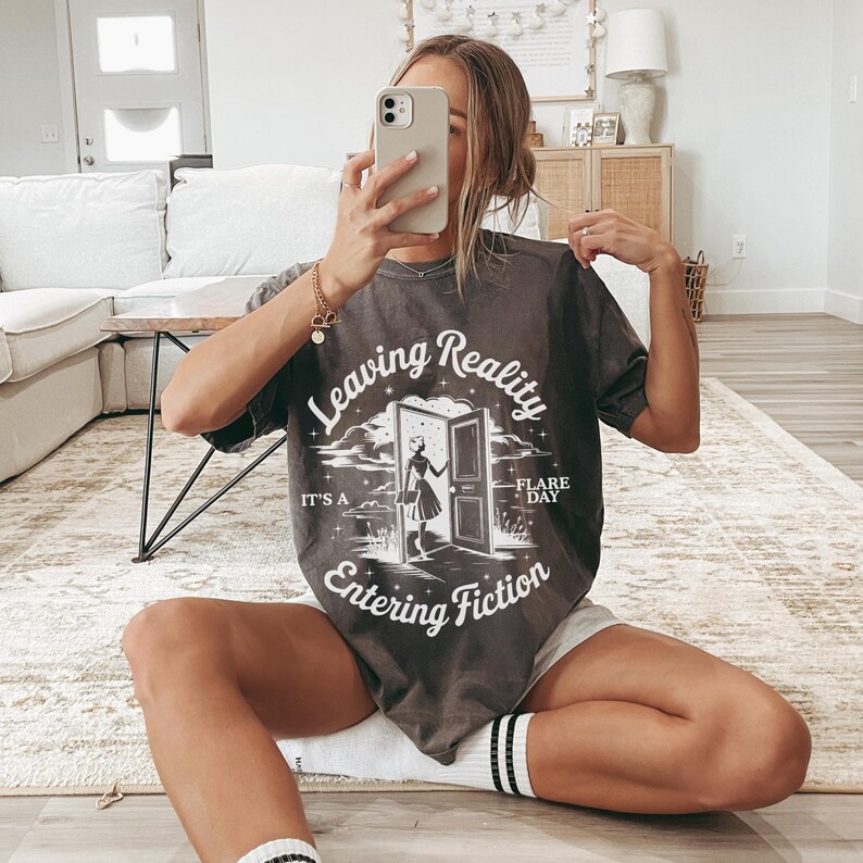 K&ouml;nnte beinhalten: Dunkelgraues T-Shirt mit dem Text "Leaving Reality Entering Fiction" und einer Grafik einer Frau an einer T&uuml;r. Das Shirt hat kurze &Auml;rmel und einen Rundhalsausschnitt. Der Text "It's a Flare Day" steht ebenfalls auf dem Shirt.