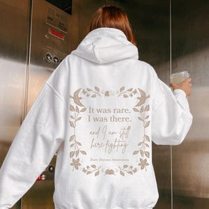 May include: White hooded sweatshirt with a floral design and the text "It was rare. I was there. and I am still here fighting. Rare Disease Awareness."