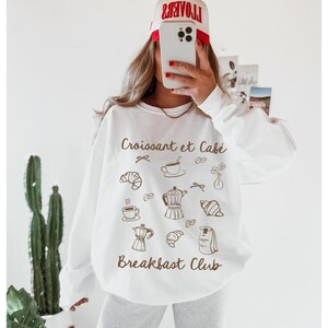 May include: White sweatshirt with a brown graphic print of croissants, coffee cups, and coffee pots. The text "Croissant et Caf&eacute;" and "Breakfast Club" is printed on the sweatshirt.