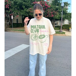 以下が含まれることがあります： 抹茶クラブ愛好家と書いてある、薄いベージュのTシャツ。緑とピンクのグラフィックデザインで、泡立て器のついた抹茶のカップと、普通の抹茶またはアイス抹茶のカップが描かれている。