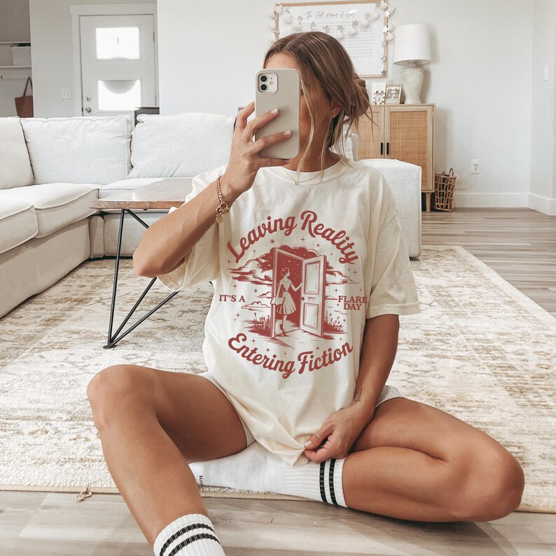 K&ouml;nnte beinhalten: Cremefarbenes T-Shirt mit dem Text "Leaving Reality Entering Fiction" in Rot. Die Grafik zeigt eine Person, die durch eine T&uuml;r geht. Das Shirt wird mit wei&szlig;en Shorts und schwarz-wei&szlig; gestreiften Socken getragen.