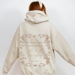 May include: A beige hoodie with a floral design and the text "It was rare. I was there. and I am still here fighting. Rare Disease Awareness" printed on the back.