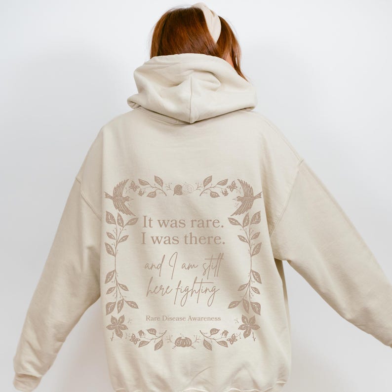 May include: A beige hoodie with a floral design and the text "It was rare. I was there. and I am still here fighting. Rare Disease Awareness" printed on the back.