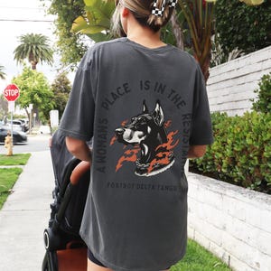 K&ouml;nnte beinhalten: Ein dunkelgraues T-Shirt mit einem Grafikdruck eines schwarzen Dobermanns mit Flammen und einer Kette um den Hals. Der Text "A woman's place is in the resistance" ist in einem Kreis um den Hund geschrieben. Der Text "Fox Trot Delta Tango" steht unter dem Grafikdruck.