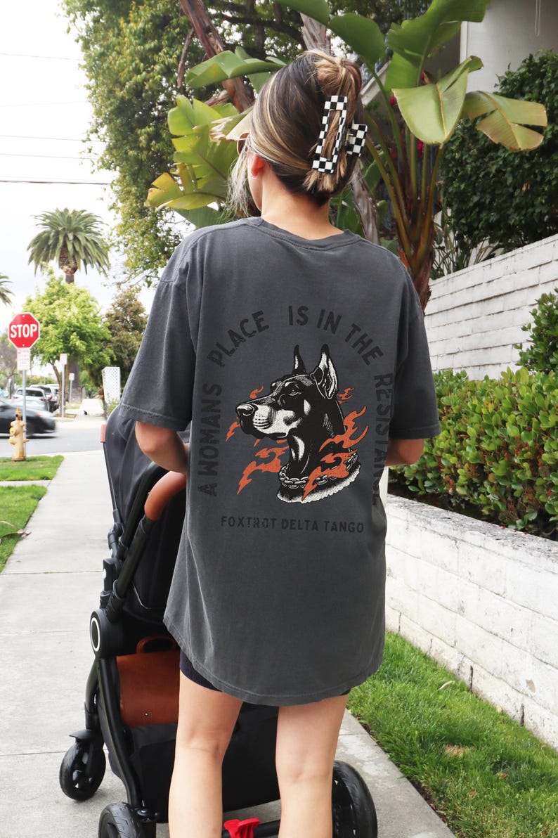 K&ouml;nnte beinhalten: Ein dunkelgraues T-Shirt mit einem Grafikdruck eines schwarzen Dobermanns mit Flammen und einer Kette um den Hals. Der Text "A woman's place is in the resistance" ist in einem Kreis um den Hund geschrieben. Der Text "Fox Trot Delta Tango" steht unter dem Grafikdruck.