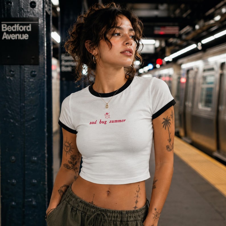 May include: A white ringer t-shirt with black trim on the collar and sleeves. The front features a small red graphic of a ladybug above the red text "sad bug summer" centered on the chest. The shirt has a cropped fit and a simple crew neckline.