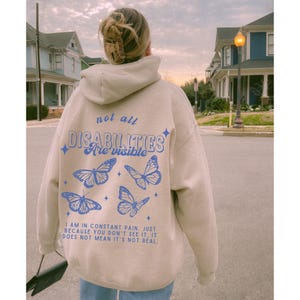 Pu&ograve; includere: Una felpa con cappuccio beige con un design a farfalla blu e il testo "not all disabilities are visible" e "I am in constant pain. Just because you don't see it, it does not mean it's not real."