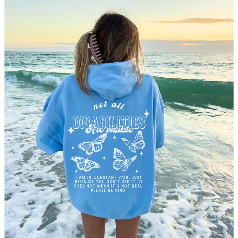 K&ouml;nnte beinhalten: Hellblauer Kapuzenpullover mit dem Text "not all DISABILITIES are visible" und einer Botschaft &uuml;ber unsichtbare Behinderungen. Der Pullover hat ein Schmetterlingsdesign.