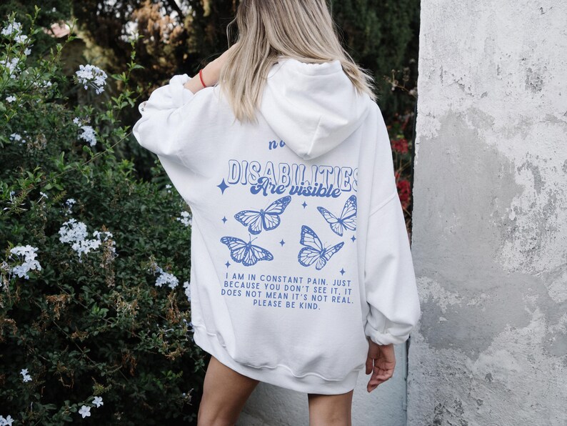 K&ouml;nnte beinhalten: Wei&szlig;er Hoodie mit einem blauen Schmetterlings-Motiv und dem Text "no disabilities are visible". Der Text unter dem Motiv lautet "I am in constant pain. Just because you don't see it, it does not mean it's not real. Please be kind."