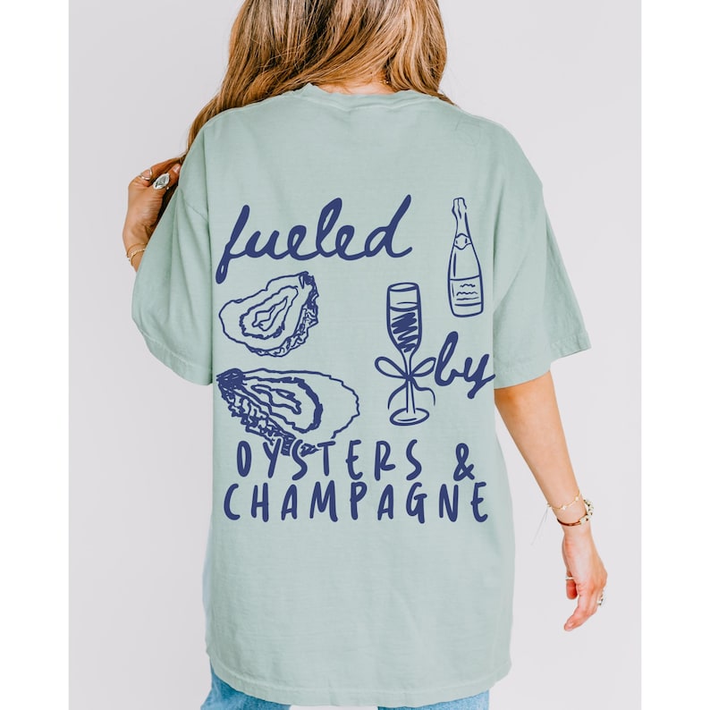 Puede incluir: Una camiseta verde claro con un gr&aacute;fico azul que dice "fueled by oysters & champagne". El gr&aacute;fico tambi&eacute;n incluye dibujos de ostras y una copa de champ&aacute;n.