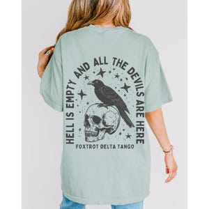 May include: Sage green t-shirt with a graphic design on the back. The design features a black raven perched on a skull, surrounded by stars, and the text "HELL IS EMPTY AND ALL THE DEVILS ARE HERE." The text "FOXTROT DELTA TANGO" is below the skull.