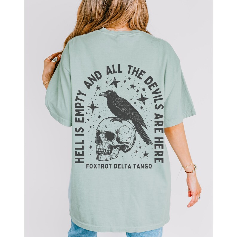 May include: Sage green t-shirt with a graphic design on the back. The design features a black raven perched on a skull, surrounded by stars, and the text "HELL IS EMPTY AND ALL THE DEVILS ARE HERE." The text "FOXTROT DELTA TANGO" is below the skull.