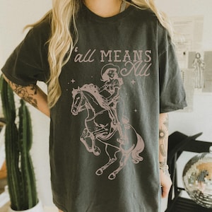 May include: Dark grey t-shirt featuring a pink graphic of a cowgirl riding a horse, with the text "Y'all Means All" in a matching pink hue. The design includes stars and a vintage aesthetic. The shirt is a casual, unisex style.