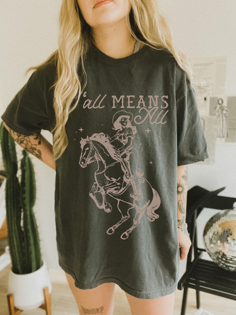 May include: Dark grey t-shirt featuring a pink graphic of a cowgirl riding a horse, with the text "Y'all Means All" in a matching pink hue. The design includes stars and a vintage aesthetic. The shirt is a casual, unisex style.
