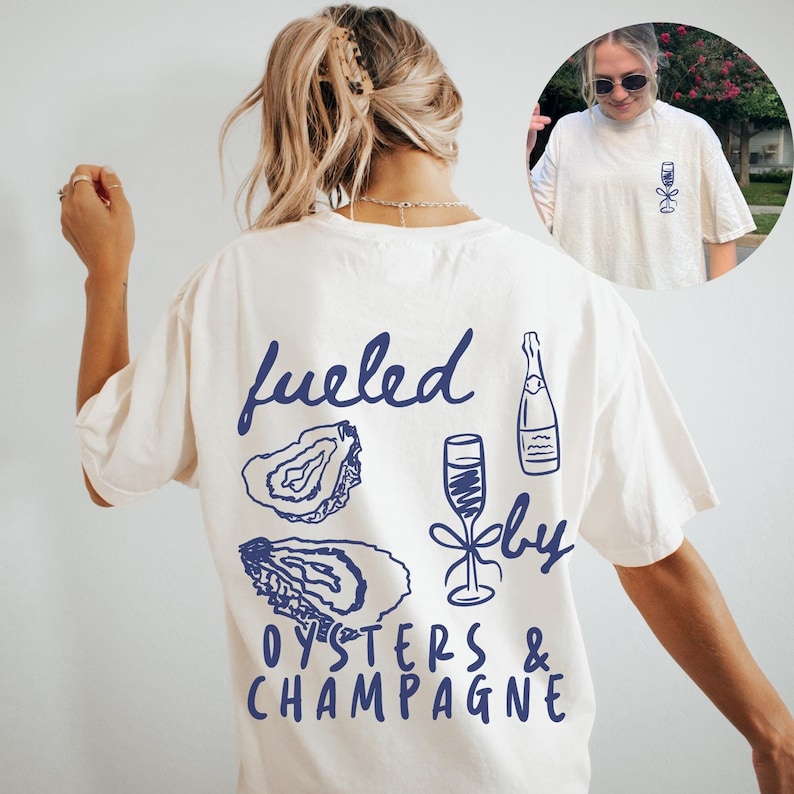 Puede incluir: Camiseta blanca con un gr&aacute;fico azul que dice "fueled by oysters & champagne" con ilustraciones de ostras, una botella de champ&aacute;n y una copa de champ&aacute;n.