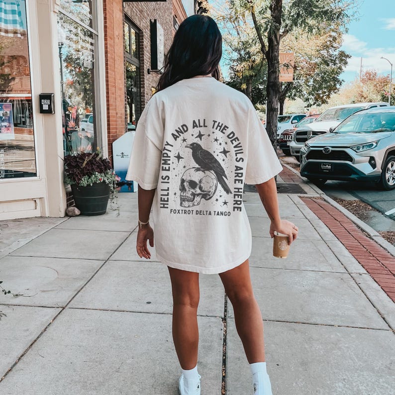 May include: Cream-colored oversized t-shirt with a graphic on the back featuring a raven perched on a skull, surrounded by stars and the text "HELL IS EMPTY AND ALL THE DEVILS ARE HERE". The shirt is worn outdoors.