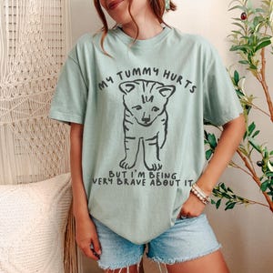 Puede incluir: Camiseta verde claro con el texto "MY TUMMY HURTS" sobre una ilustraci&oacute;n negra de un cachorro. Debajo, el texto dice "BUT I'M BEING VERY BRAVE ABOUT IT."