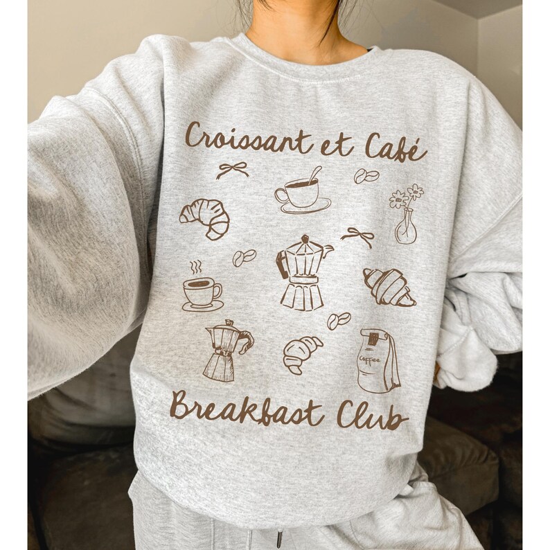 May include: Gray sweatshirt with the text "Croissant et Caf&eacute; Breakfast Club" in brown lettering. The design includes illustrations of croissants, coffee cups, coffee beans, and a coffee pot.