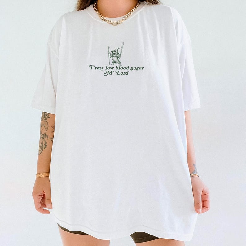 May include: White t-shirt with a green graphic of a wizard and the text "T'was low blood sugar M' Lord". The shirt has short sleeves and a relaxed fit. The shirt is made of a soft, comfortable material.