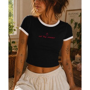May include: A black ringer t-shirt with white trim on the sleeves and neckline. The chest features a small red graphic of a ladybug above the red text "sad bug summer" written in a lowercase, cursive font. The shirt has a cropped fit.