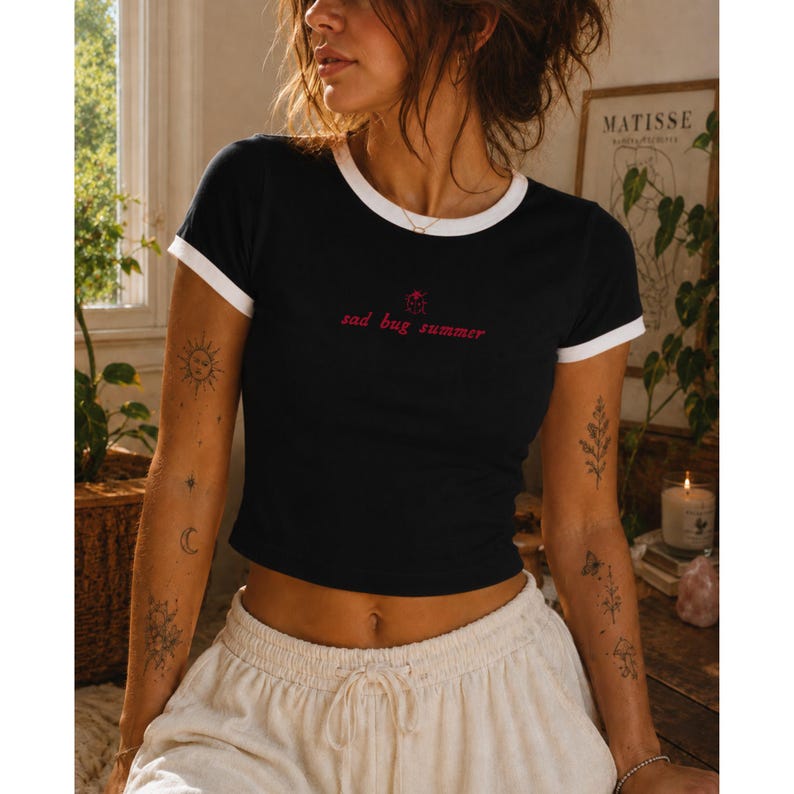 May include: A black ringer t-shirt with white trim on the sleeves and neckline. The chest features a small red graphic of a ladybug above the red text "sad bug summer" written in a lowercase, cursive font. The shirt has a cropped fit.