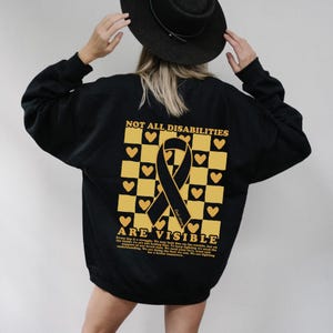 May include: Black sweatshirt with a yellow and black checkered pattern and a black ribbon. The text "Not All Disabilities Are Visible" is printed on the sweatshirt.