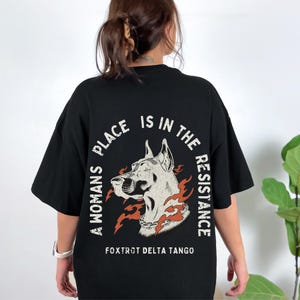 K&ouml;nnte beinhalten: Schwarzes T-Shirt mit einem Grafikdruck eines Hundekopfes mit Flammen und dem Text "A woman's place is in the resistance" und "Foxtrot Delta Tango"