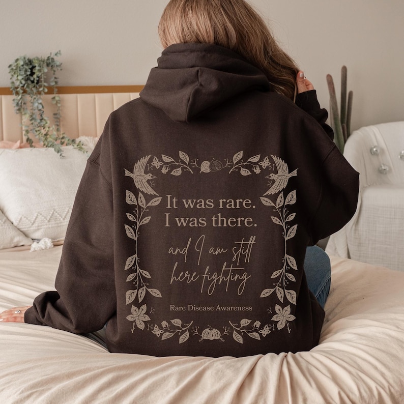May include: A brown hooded sweatshirt with a floral design and the text "It was rare. I was there. and I am still here fighting. Rare Disease Awareness."