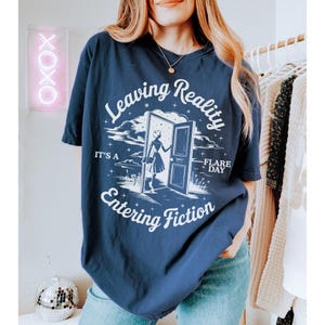 K&ouml;nnte beinhalten: Marineblaues T-Shirt mit dem Text "Leaving Reality Entering Fiction" in einer verspielten Schriftart. Das Design zeigt eine Frau, die durch eine T&uuml;r geht. Das Shirt ist ein bequemer, l&auml;ssiger Stil.