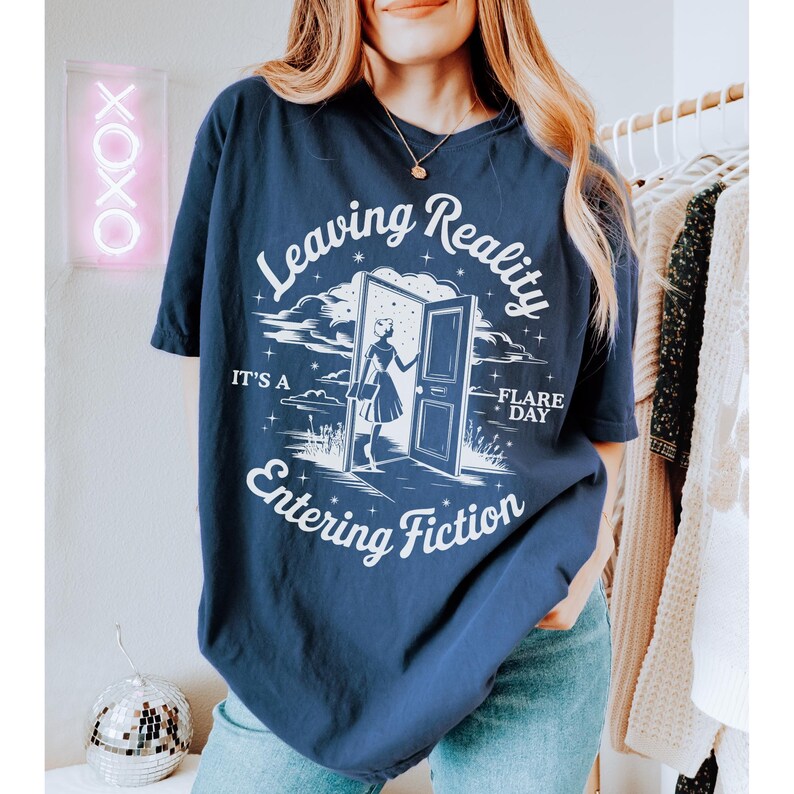 K&ouml;nnte beinhalten: Marineblaues T-Shirt mit dem Text "Leaving Reality Entering Fiction" in einer verspielten Schriftart. Das Design zeigt eine Frau, die durch eine T&uuml;r geht. Das Shirt ist ein bequemer, l&auml;ssiger Stil.