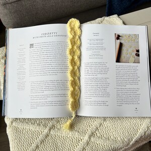 May include: A close-up of an open cookbook with a yellow crochet bookmark. The page shows a recipe for Corzetti with Pesto alla Genovese. The recipe includes instructions for making the pasta dough and shaping the corzetti.