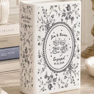 May include: A white ceramic book-shaped vase featuring a black floral toile pattern. The front displays an oval frame with the text "Joe & Karen", "Engaged", and "05.15.2026" alongside a graphic of two clinking champagne glasses with ribbons.