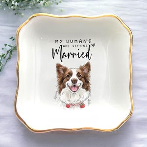 May include: A white ceramic trinket dish with a gold rim and a scalloped edge. The dish features a smiling dog illustration and the text "MY HUMANS ARE GETTING Married". A charming wedding gift.